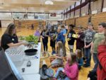 Hydroponics Youth Event as children sit on the gym floor and look at a presentation at a table